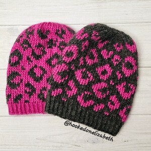 May include: Two knitted hats with a leopard print pattern. One hat is pink with black spots and the other is gray with pink spots. The hats are on a white wooden surface. @hookedonelizabeth