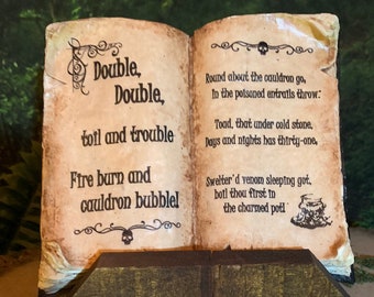 May include: An open book ornament with aged pages, displaying spell text. The book rests on a dark wooden stand. The text includes phrases such as "Double, toil and trouble" and "Fire burn and cauldron bubble!" A decorative item.