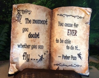 May include: A decorative open book with a weathered appearance. The pages display the quote "The moment you doubt whether you can fly..." and "You cease for EVER to be able to do it... ~ Peter Pan."