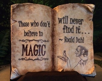 May include: An open book with aged paper pages, featuring the quote "Those who don't believe in magic will never find it... ~ Roald Dahl." The book has a dark brown base and is set against a blurred green background.