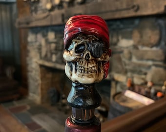 May include: A skull-shaped bottle stopper with a red bandana and an eye patch. The skull is detailed with a black base and sits atop a wine bottle. The bottle has a red foil top and a white label.