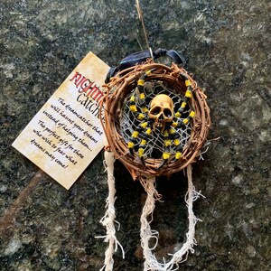 May include: A handmade dreamcatcher featuring a skull spider. The spider has yellow and black stripes. The dreamcatcher is made of woven twigs and white string. A tag reads "Nightmare Catchers".