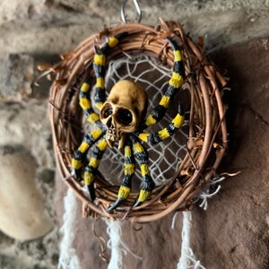 May include: A Halloween dream catcher with a skull-faced spider. The spider has yellow and black striped legs and is set in a woven wreath with a white web and hanging white yarn. The background is a stone wall.