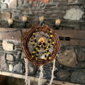 May include: A Halloween decoration featuring a spider with a skull-shaped head and yellow and black striped legs. The spider is set in a woven wreath with white string accents, suspended by a silver wire. Ideal for seasonal decor.