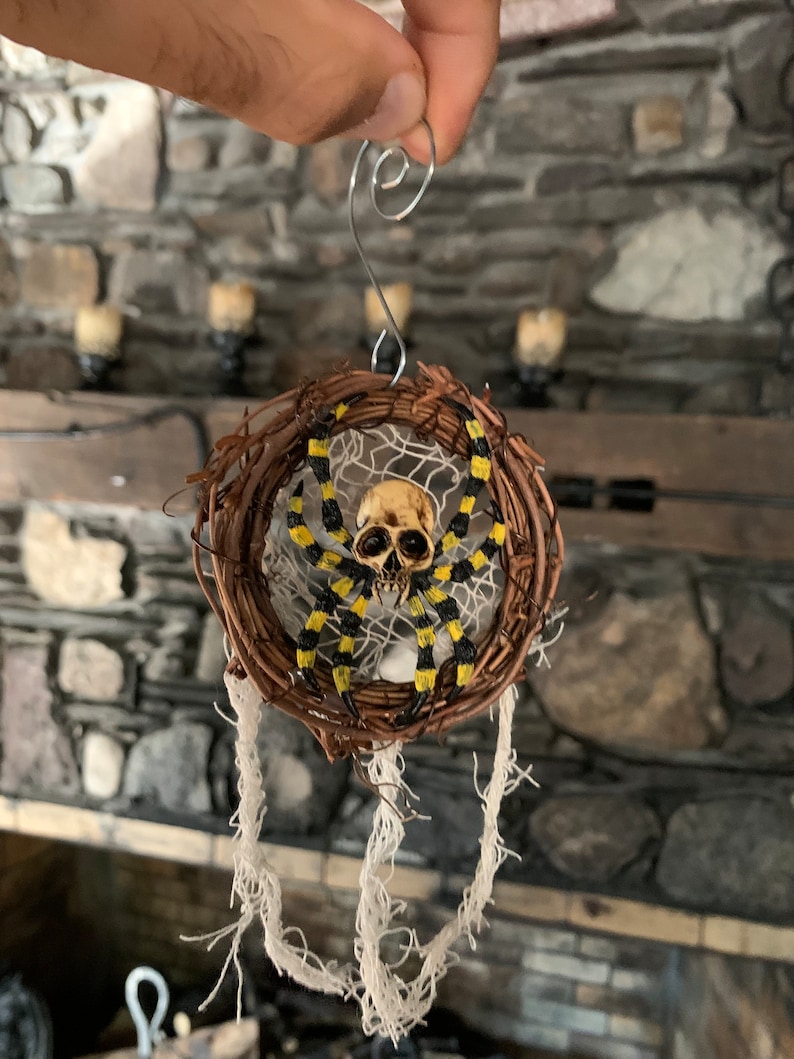May include: A Halloween decoration featuring a spider with a skull-shaped head and yellow and black striped legs. The spider is set in a woven wreath with white string accents, suspended by a silver wire. Ideal for seasonal decor.