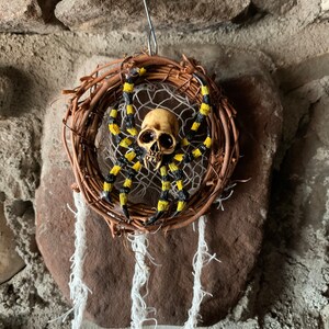 May include: A Halloween dreamcatcher with a skull-shaped centrepiece and a spider with yellow and black striped legs. The dreamcatcher is made of woven twigs and has white hanging strands.