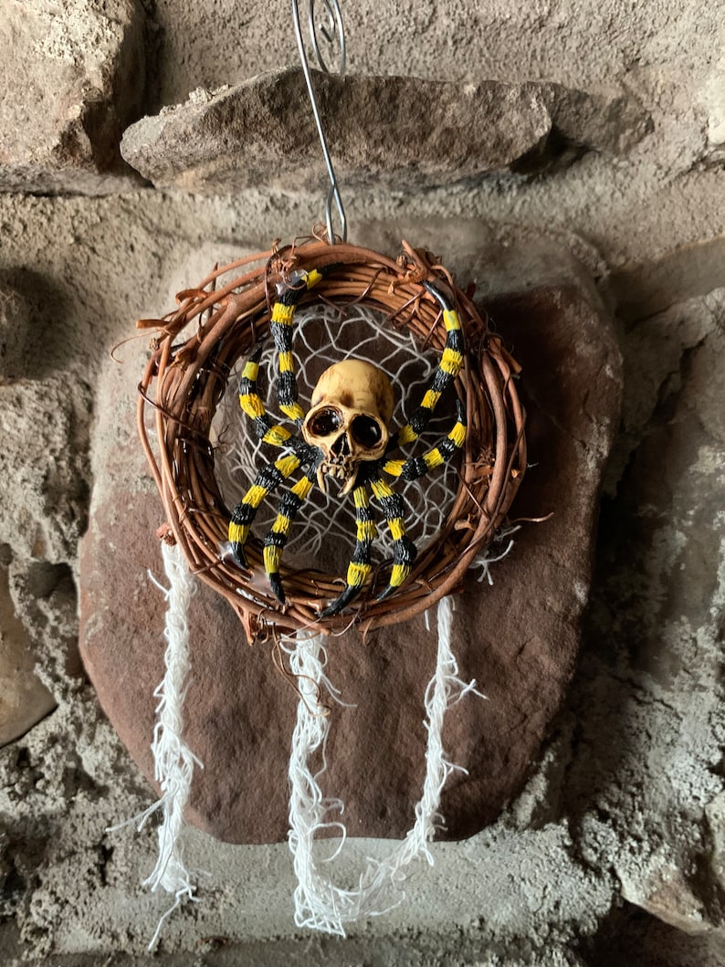 May include: A Halloween dreamcatcher with a skull-shaped centrepiece and a spider with yellow and black striped legs. The dreamcatcher is made of woven twigs and has white hanging strands.