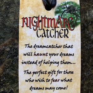 May include: A rectangular tan tag with the words "Nightmare Catcher" in red and black lettering. The tag has a black ribbon and a small image of a spider. The text on the tag reads: "The dreamcatcher that will haunt your dreams instead of helping them..."