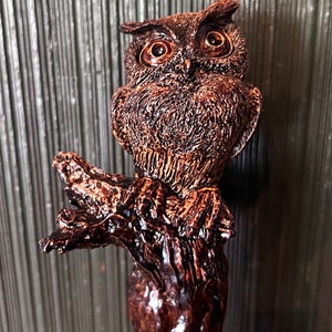 May include: A detailed, brown owl figurine perched on a textured branch. The owl has large, expressive eyes and intricate feather detailing. The figurine is made of a glossy, dark brown material, giving it a rustic appearance. The overall design is artistic and decorative.