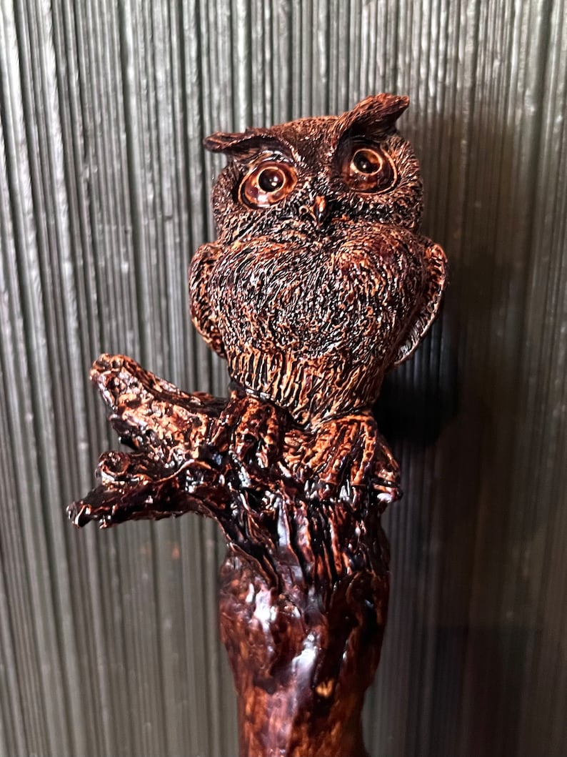 May include: A detailed, brown owl figurine perched on a textured branch. The owl has large, expressive eyes and intricate feather detailing. The figurine is made of a glossy, dark brown material, giving it a rustic appearance. The overall design is artistic and decorative.