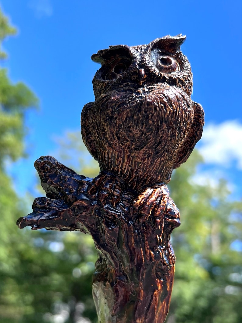 May include: A detailed, brown owl sculpture perched on a wooden branch. The owl has textured feathers and large, expressive eyes. The wood has a dark, glossy finish. The background is a bright blue sky with some clouds.
