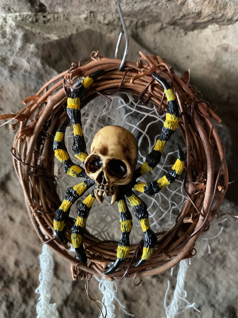 May include: A Halloween dream catcher. It features a skull-shaped spider with yellow and black striped legs, set within a woven wreath. A white web and hanging strands complete the design.