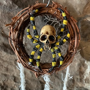 May include: A Halloween decoration featuring a spider with a skull-shaped head and striped yellow and black legs. The spider is mounted on a woven wreath with hanging white strands, creating a spooky aesthetic.