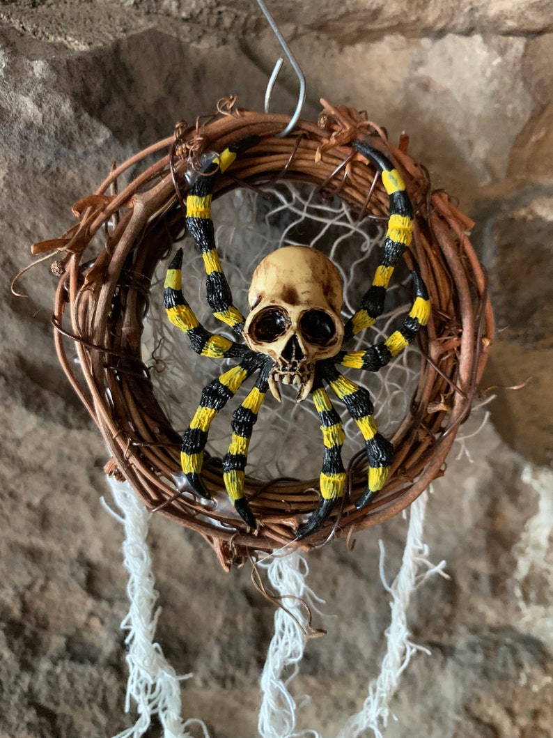 May include: A Halloween decoration featuring a spider with a skull-shaped head and striped yellow and black legs. The spider is mounted on a woven wreath with hanging white strands, creating a spooky aesthetic.