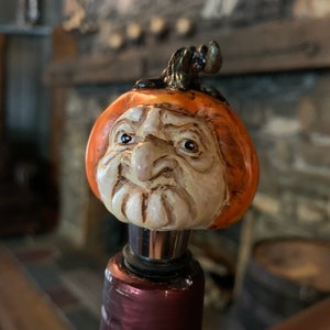 May include: A decorative bottle stopper featuring a carved pumpkin head with a detailed face and a green stem. The pumpkin is orange and cream-colored, mounted on a silver and burgundy stopper, perfect for wine bottles.