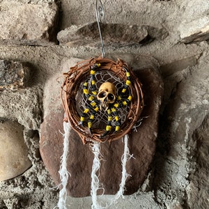 May include: A handcrafted dreamcatcher with a skull centerpiece and a woven vine frame. The dreamcatcher features black and yellow accents and white hanging threads. The decorative item is suspended by a silver wire.