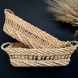 Könnte beinhalten: Zwei geflochtene Körbe aus Naturmaterialien, einer in den anderen geschachtelt. Die Körbe sind oval geformt mit einem geflochtenen Muster und haben an beiden Enden Griffe.