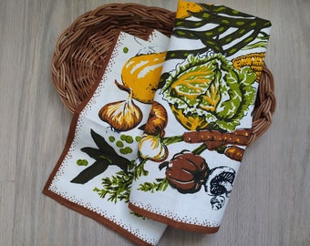 Pode incluir: Dois panos de cozinha brancos com borda marrom e estampa de vegetais. Um pano apresenta uma cebola amarela, alho e ervilhas verdes. O outro pano tem uma couve, cenouras e cogumelos. Os panos estão colocados em uma cesta de vime marrom.