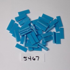 May include: A collection of small, rectangular, turquoise-colored mosaic tiles scattered on a white surface. The tiles are glossy and have a slightly rounded edge. A small white label with the number "5467" is visible.