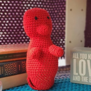 May include: A red crocheted amigurumi toy shaped like a person. The toy has black eyes and a small nose. The toy is sitting on a blue surface.