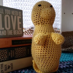 以下が含まれることがあります： 黒い目をした黄色の編みぐるみ。編みぐるみは、「All you need is love and the beach」と書かれた本が積まれた上に座っている。