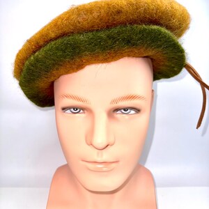 May include: A two-tone beret in gold and olive green. The hat is made of a textured, felt-like material and is displayed on a mannequin head. A brown leather cord is attached to the side of the hat.