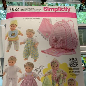 May include: A Simplicity sewing pattern for 15-inch baby doll clothes. The pattern includes a pink and white striped backpack, a pink and white striped dress, a pink and white striped shirt, a pink and white striped pants, a green and white striped sweater, a green and white striped hat, and a green and white striped blanket. The pattern also includes a QR code for free instructions on knit and crochet doll accessories.