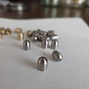 May include: A collection of metallic, bullet-shaped fishing weights. Some are silver-colored, while others have a brass or gold finish. The weights are arranged on a white surface, with a blurred background.