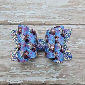 May include: A large, handmade hair bow featuring a blue and purple pattern with images of animated characters. The bow has glitter accents on the edges and center, and is set against a light wood background.