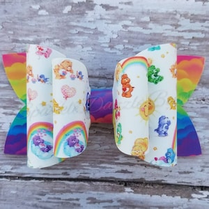 May include: A large, handmade hair bow featuring a colorful rainbow and Care Bears pattern. The bow has a white base with various Care Bear characters, rainbows, and hearts. The edges are a vibrant rainbow gradient.