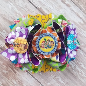 May include: A large, multi-layered hair bow featuring various colorful ribbons. The bow includes purple polka dot, green, and patterned ribbons. A central circular embellishment showcases a Toy Story character design, surrounded by rhinestones.