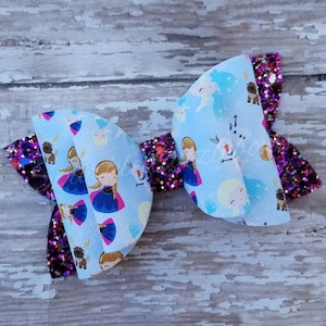 May include: A handmade hair bow featuring a light blue print with cartoon characters and a glittery purple accent. The bow has a layered design with a central knot and is suitable for children's accessories.