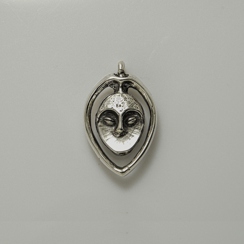 May include: Silver pendant featuring a stylized face with closed eyes, set within a teardrop-shaped frame.