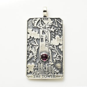 May include: Silver pendant featuring the Tower card from the tarot deck. The pendant is engraved with the image of a tower being struck by lightning, with people falling from the tower. The pendant has a purple gemstone in the center and the words "THE TOWER" at the bottom.