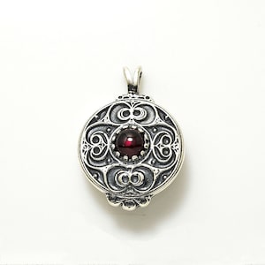 May include: A silver pendant with an intricate design featuring a red gemstone in the center. The pendant is round and has a detailed filigree pattern.