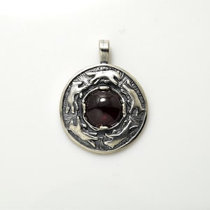 May include: A silver pendant featuring a dark red gemstone at its centre, encircled by five stylised animal figures. The pendant is displayed against a white backdrop.