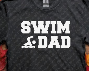 May include: Black t-shirt with the words "SWIM DAD" in large white letters, along with a swimmer graphic. The shirt is displayed on a wooden surface with colorful autumn leaves scattered around it.