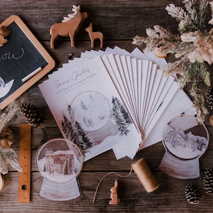 May include: A flat lay of a craft project with a variety of winter-themed items, including a chalkboard with the word "Snow" written on it, a stack of cards with the title "Snow Globes", two snow globes, a wooden reindeer, a wooden squirrel, a wooden rabbit, a wooden house, pine cones, and a ruler.