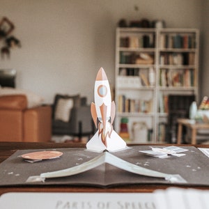 May include: A pop-up book with a rocket ship, planets, and a space shuttle. The book is open on a wooden table with a notebook that says "Parts of Speech" on the cover.
