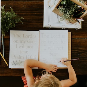May include: A child is writing on a worksheet with the text "The Lord is on my side; I will not fear. Psalm 118:6". A wooden box with colored pencils and greenery sits nearby. The scene is set on a dark wooden table.