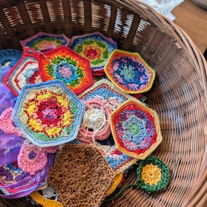 May include: A wicker basket filled with colorful, handmade crochet pieces. The pieces are various shapes, including hexagons and circles, and feature vibrant rainbow colors. The basket is brown and the crochet pieces are in various stages of completion.