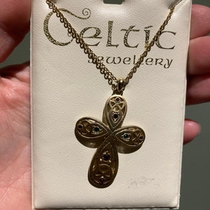May include: A gold Celtic cross necklace with green, red, and purple gemstones. The necklace is displayed on a white card with the text "Celtic Jewellery".