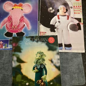 May include: Three knitting pattern books. One features a pink elephant-like creature with the text "HELLO, TINY!". Another shows an astronaut with the text "TO BOLDLY GO". The third depicts a green figure with the text "GREEN MAN".