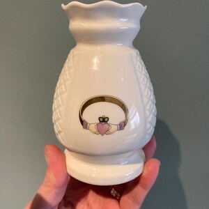 Peut inclure: Un vase en céramique blanche avec un motif tissé et un nœud celtique représentant un cœur et une couronne. Le vase a un bord festonné et une petite base.