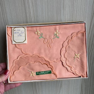 Könnte beinhalten: Ein Set aus drei pfirsichfarbenen Leinen-Taschentüchern mit gewellten Rändern und gestickten Blumen. Die Taschentücher sind in einer weißen Schachtel mit einem grünen Etikett verpackt, auf dem "All Pure Linen Made in Ireland" steht.