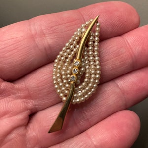 Peut inclure: Une broche en forme de feuille dorée avec une rangée de perles et trois strass transparents au centre.