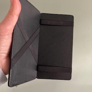 May include: Open black wallet with elastic bands and a cross-strap design. The wallet is made of a smooth, dark material and appears to be open, revealing the interior. The design suggests a minimalist approach to carrying cards and cash.