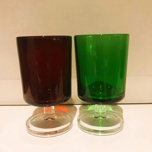 Peut inclure: Deux verres à pied avec des bols cylindriques, l'un rouge foncé et l'autre vert vif. Chaque verre présente une base circulaire transparente et un pied court et épais en verre transparent soutenant le récipient coloré, au design simple.