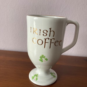 Puede incluir: Taza de cerámica blanca con una inscripción dorada que dice "Irish Coffee". La taza tiene una base con pie y está decorada con tréboles verdes.