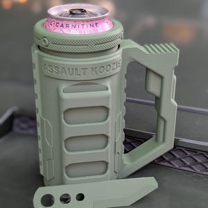 May include: A light olive green 3D-printed beverage holder with a handle and the text "ASSAULT KOOZIE" embossed on the front. The holder contains a pink and silver aluminium can. A small, matching olive green plastic knife-shaped tool lies in front.